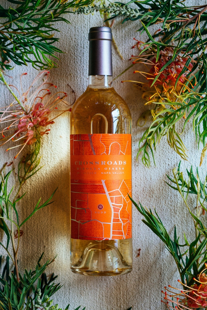 A bottle of Crossroads Ribolla Gialla surrounded by green pine branches and red flowers on a textured light background.