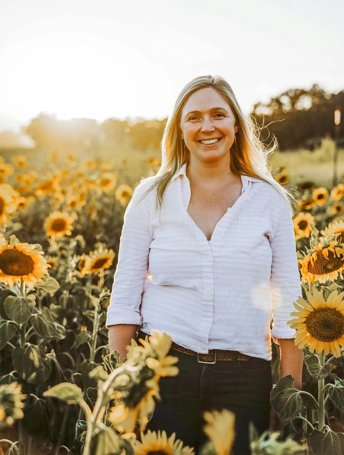 A smiling woman standing in a sunflower field at sunset, wearing a white button-up shirt and dark pants.