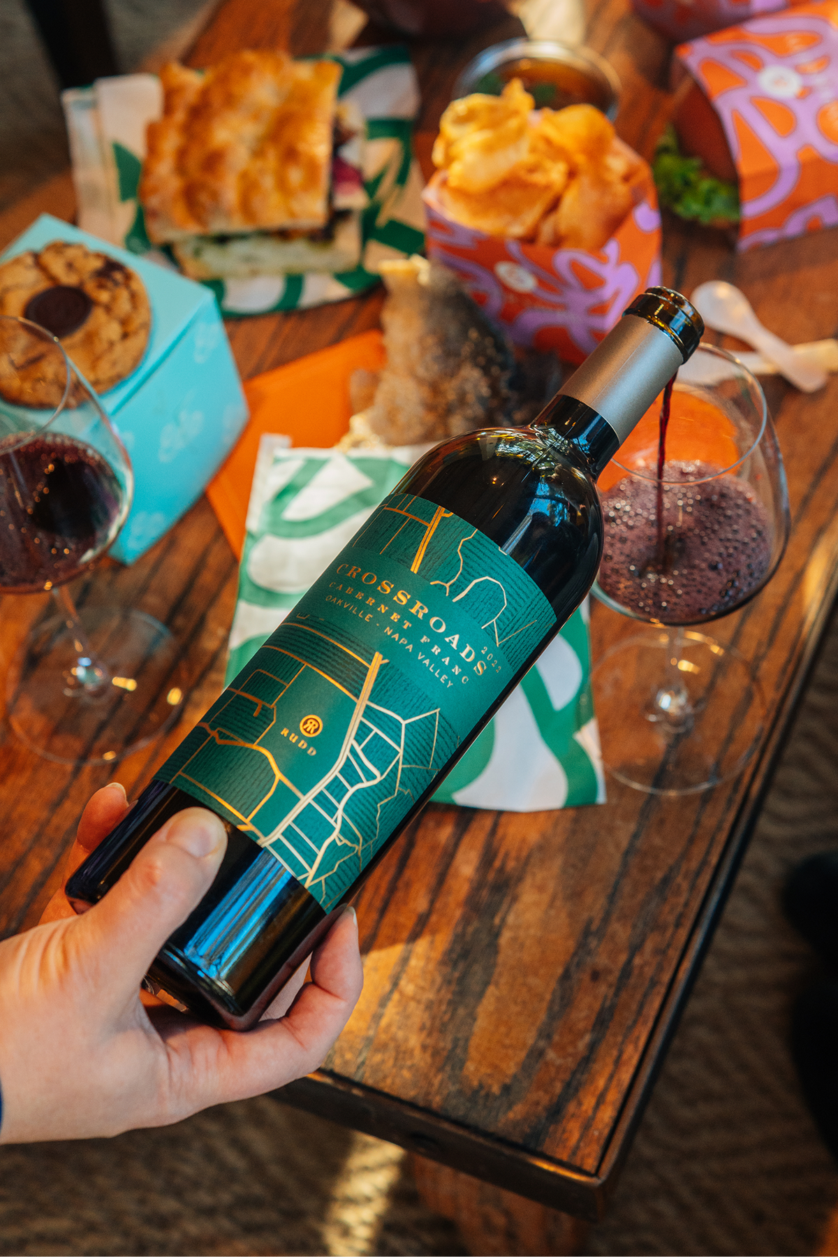 Crossroads Cabernet Franc wine bottle being poured with glasses of red wine, focaccia, pastries and snacks on a table.