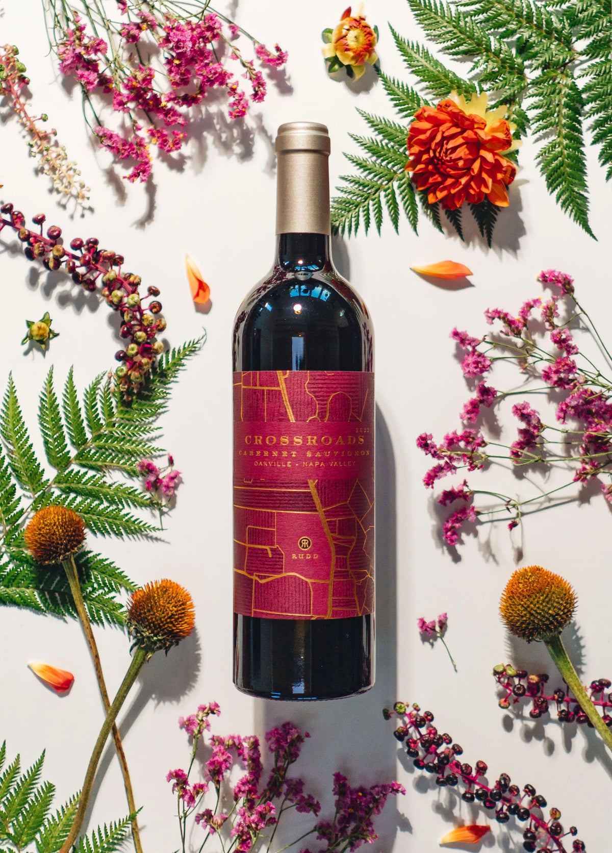A bottle of Crossroads Cabernet Sauvignon wine is centered on a white background, surrounded by decorative botanical elements including green ferns, orange flowers, and pink blossoms.