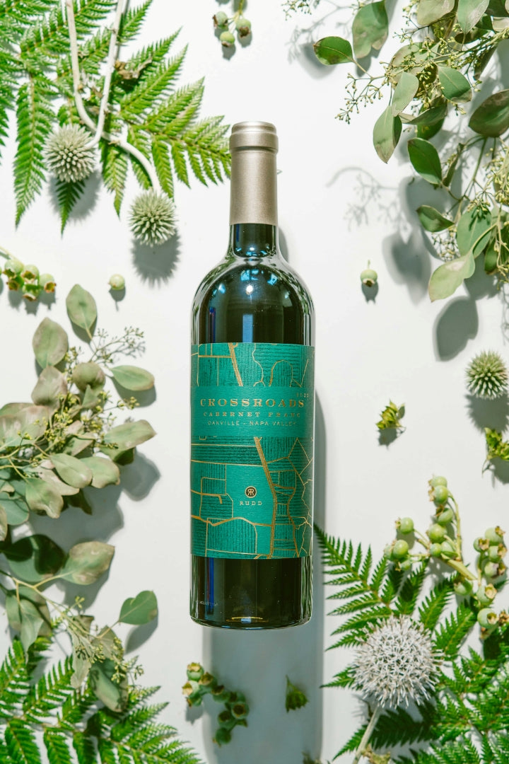 A bottle of Crossroads Cabernet Franc wine with a teal and gold label, styled in a flat lay with green ferns, eucalyptus, and thistles on a white background.