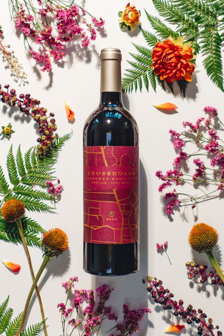 A bottle of Crossroads Cabernet Sauvignon wine centered on a white background, surrounded by decorative dried flowers, orange marigolds, and green fern leaves.