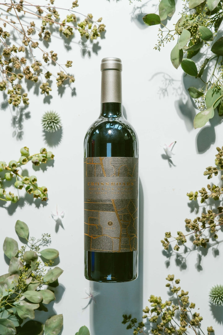 A dark glass wine bottle with a metallic gold label and silver foil cap, centered on a white background and surrounded by dried green botanicals.