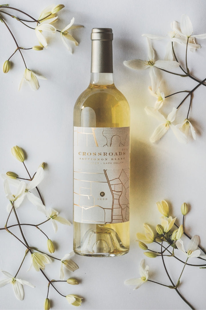A bottle of Crossroads Sauvignon Blanc white wine is centered on a white background, surrounded by delicate white flowers and green buds. The bottle has a silver foil cap and a cream-colored label with a minimalist map design.